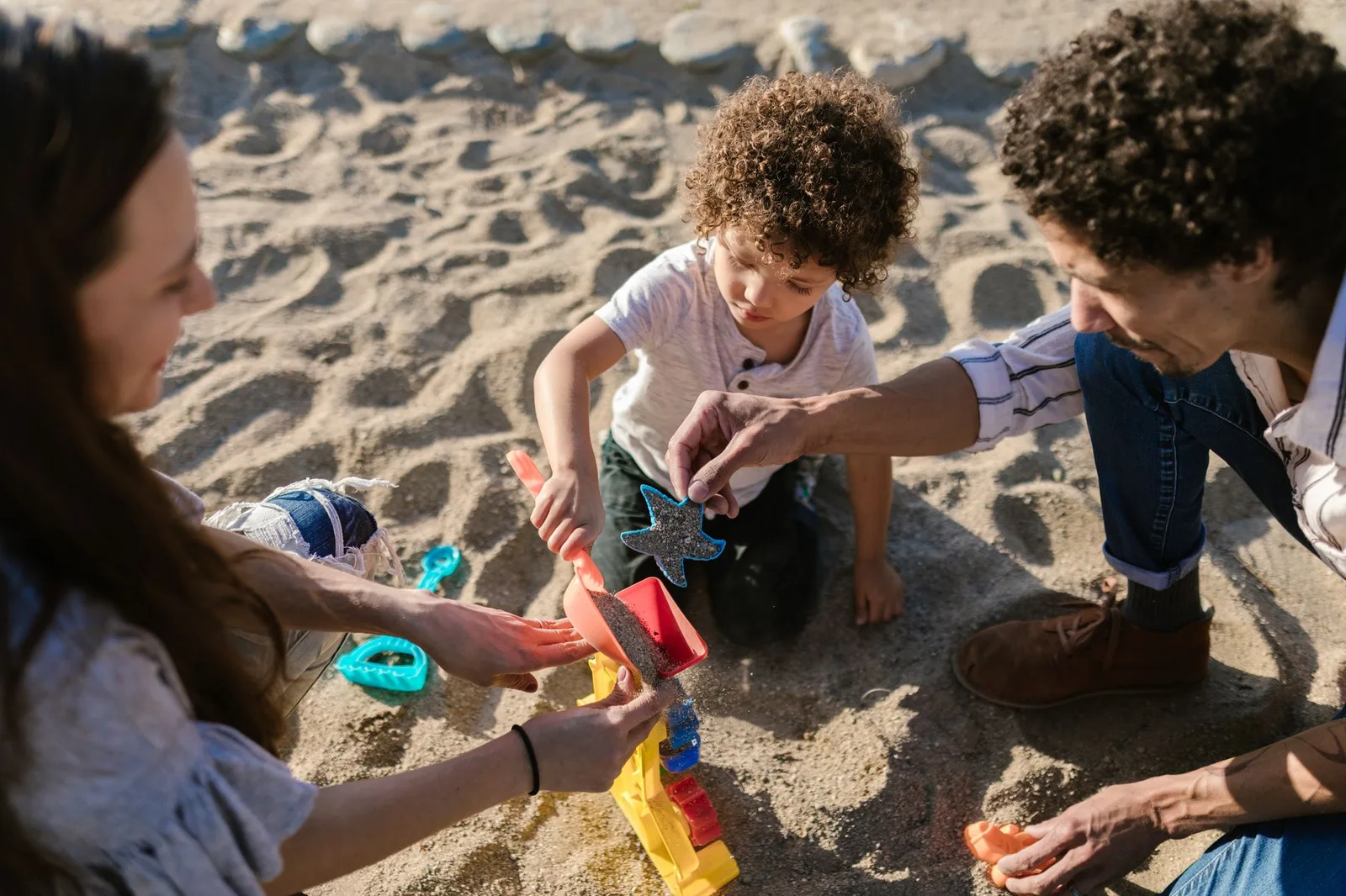 משפחה ישראלית עם שני ילדים משחקת על חוף החול הרגוע של סאנור בבאלי בשעת בוקר, עם סירות מסורתיות וים שקט ברקע