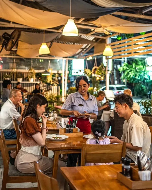 מלצרית מגישה למשפחה בשולחן עץ, מתחת ליריעות בד ומנורות תלויות בערב