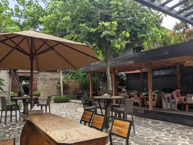 חצר מרוצפת אבן עם עץ גדול, שמשייה חומה, שולחנות עץ ואזור ישיבה מקורה
