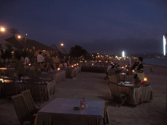 שולחנות ערוכים על החול בשעת דמדומים, נרות קטנים ואורות לאורך החוף