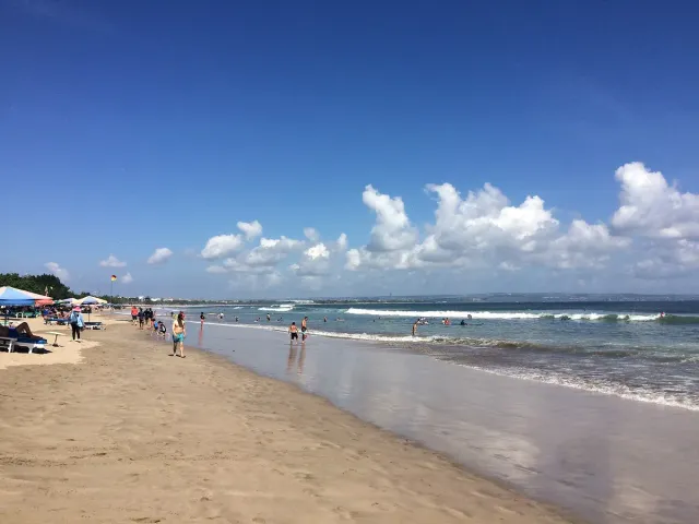 רצועת חול רחבה עם שמשיות ומיטות שיזוף, גלים נמוכים ומטיילים במים הרדודים