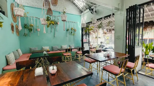 חלל עם קירות טורקיז, עציצים תלויים במקרמה, ספסלים בריפוד ורוד ושולחנות עץ